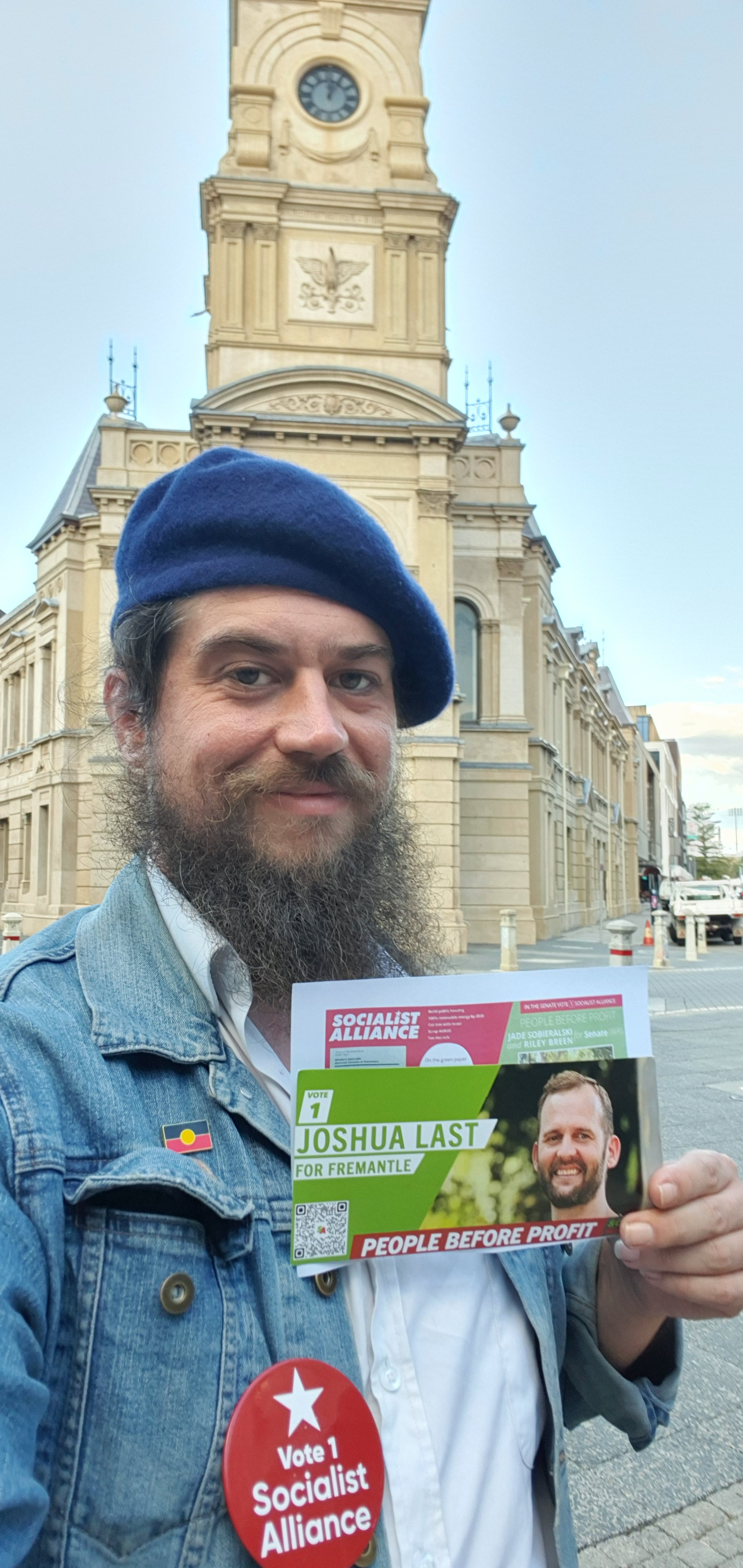 vidak standing in front of fremantle town hall. vidak is holding an election flyer that reads the name of the socialist alliance candidate JOSHUA LAST. vidak is wearing a badge that reads VOTE 1 SOCIALIST ALLIANCE. vidak is wearing an australian aboriginal flag pin. vidak standing in front of fremantle town hall. vidak is holding an election flyer that reads the name of the socialist alliance candidate JOSHUA LAST. vidak is wearing a badge that reads VOTE 1 SOCIALIST ALLIANCE. vidak is wearing an australian aboriginal flag pin.