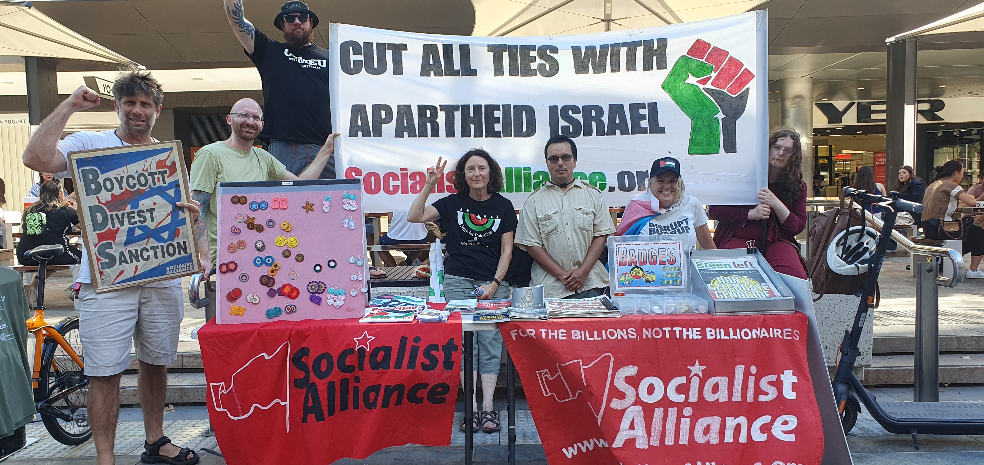 activists standing behind stalls, and in front of a banner. 

the tablecloths read "socialist alliance".

the banner reads: "CUT ALL TIES WITH APARTHEID ISRAEL"
